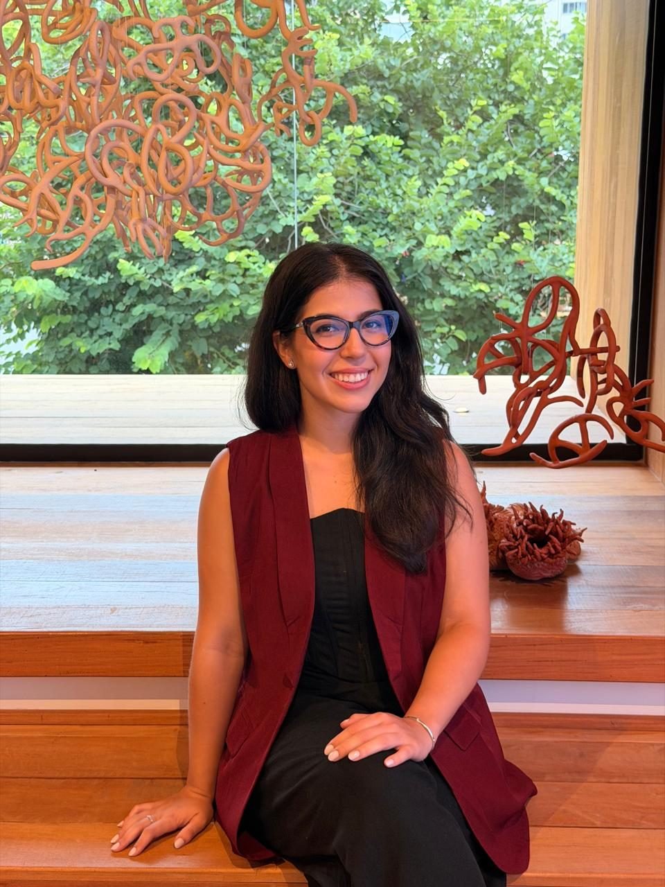 Bia, psicóloga, sorrindo, sentada num banco de madeira diante de uma janela com plantas.
