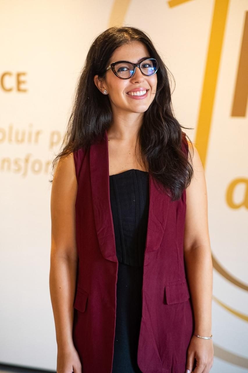 Bia em pé, sorrindo, com colete vinho, diante de uma parede clara.